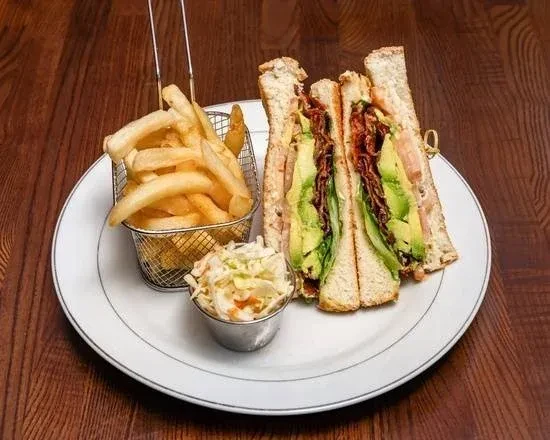 A plate with a club sandwich, French fries, and coleslaw on a wooden table.