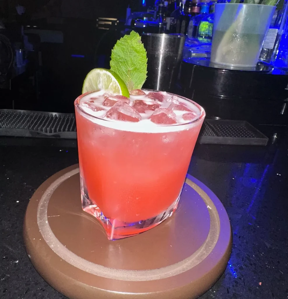 A red cocktail with lime, mint, and ice sits on a bar.