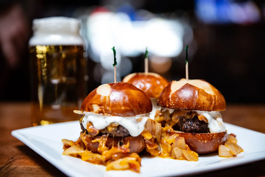 Three sliders on pretzel buns with caramelized onions and a beer.