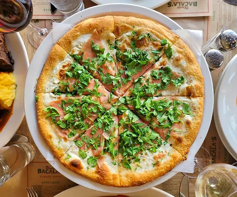 A white pizza with prosciutto and arugula, sliced on a white plate.