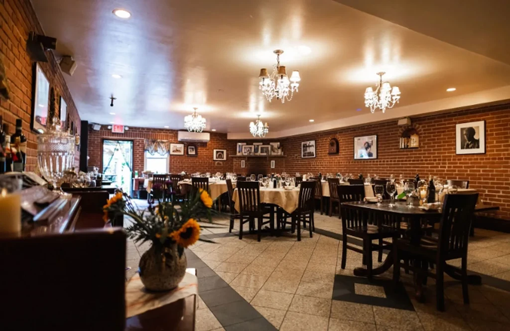 Empty restaurant dining room with brick walls, set tables, and chandeliers.