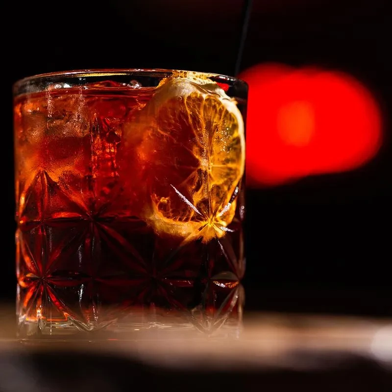 A dark red cocktail in a crystal glass with an orange slice.