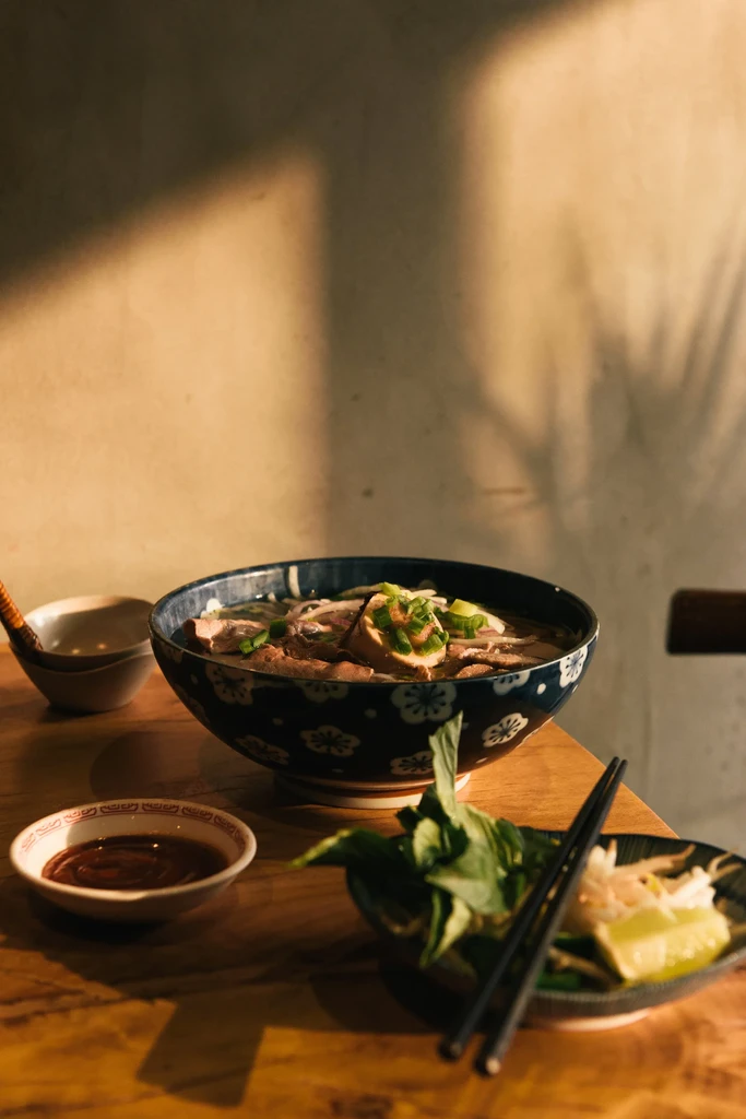 A bowl of pho with sides on a wooden table.