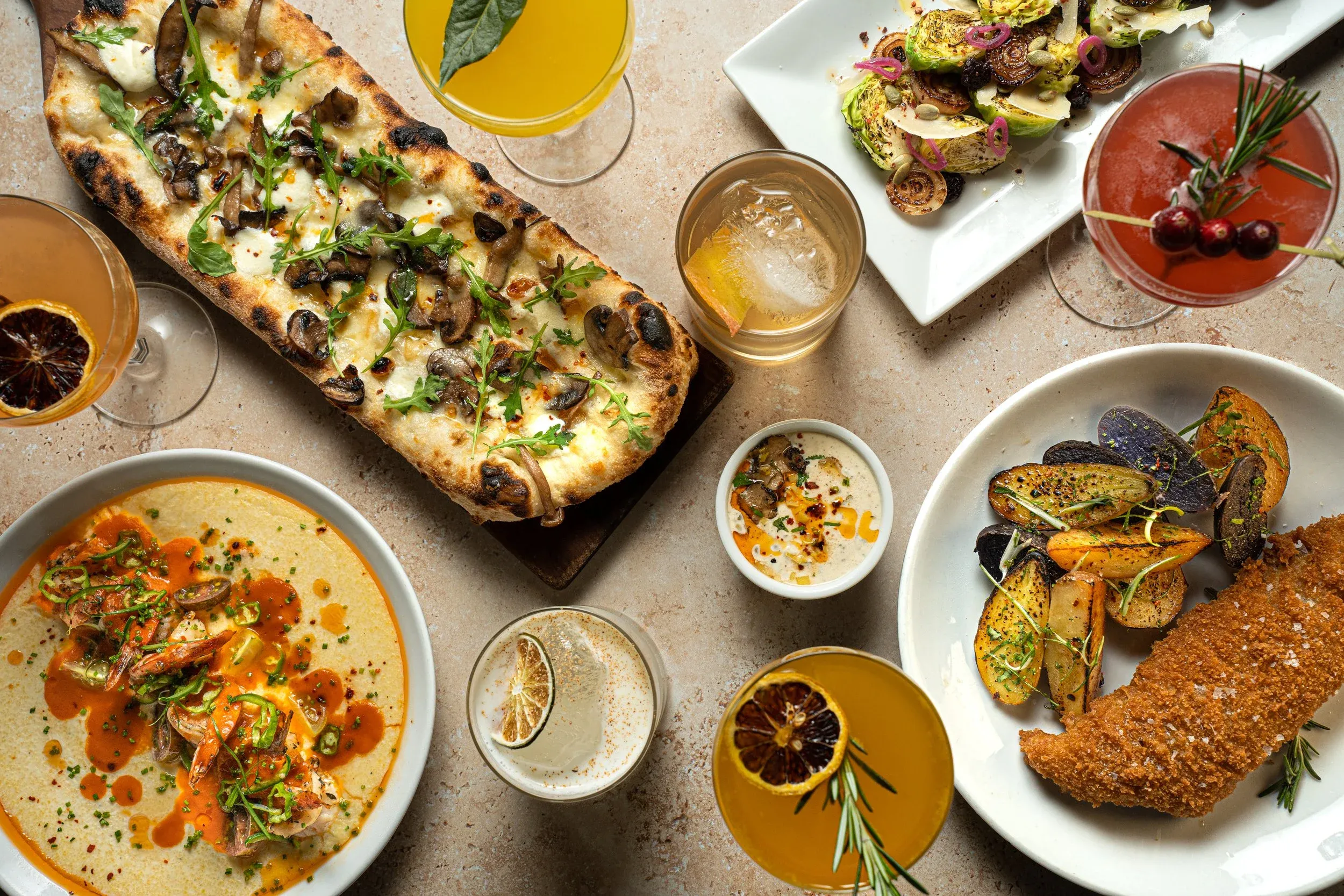 An aerial view of various dishes and cocktails on a light brown table.