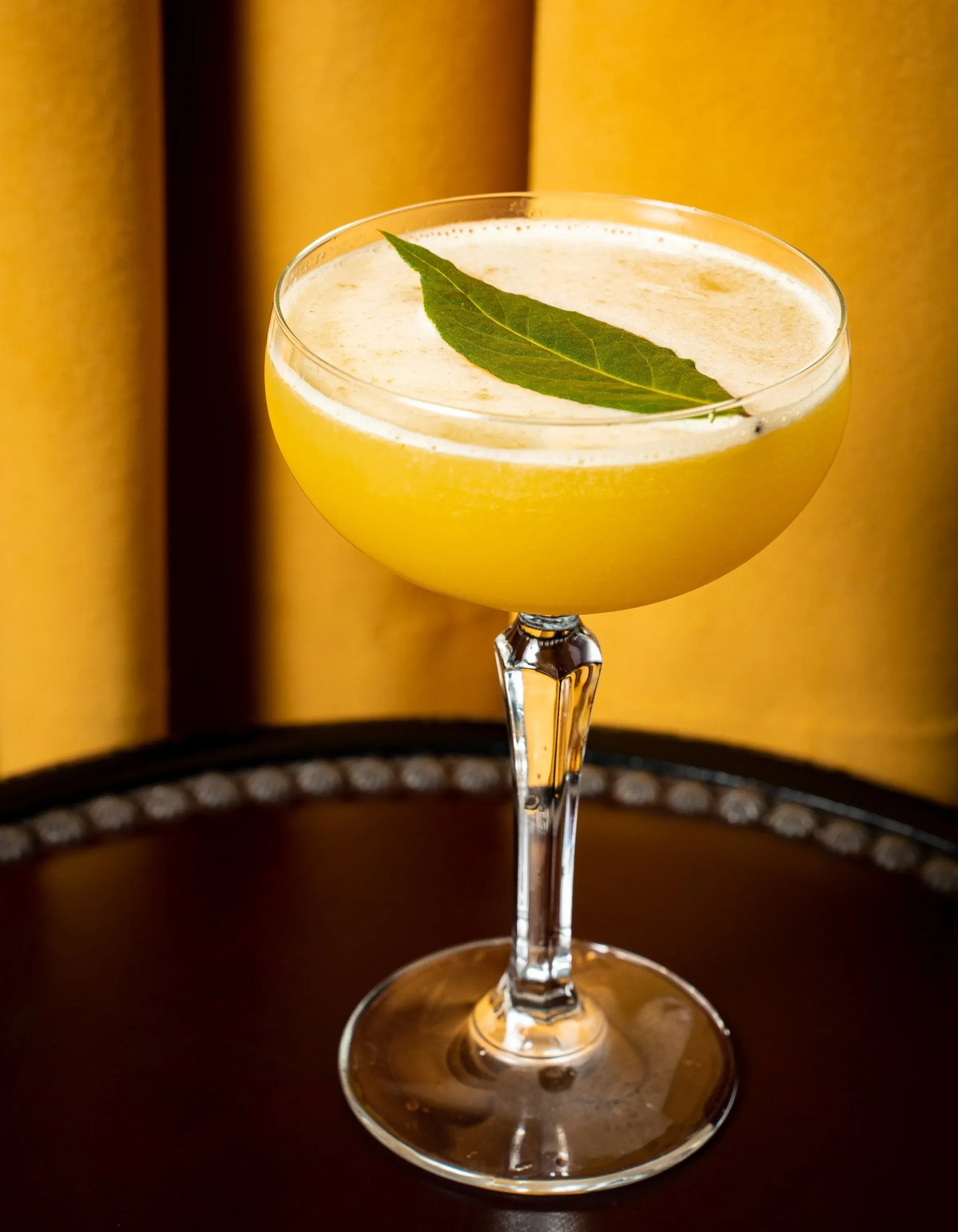 Yellow cocktail with foamed top and a single green leaf garnish.