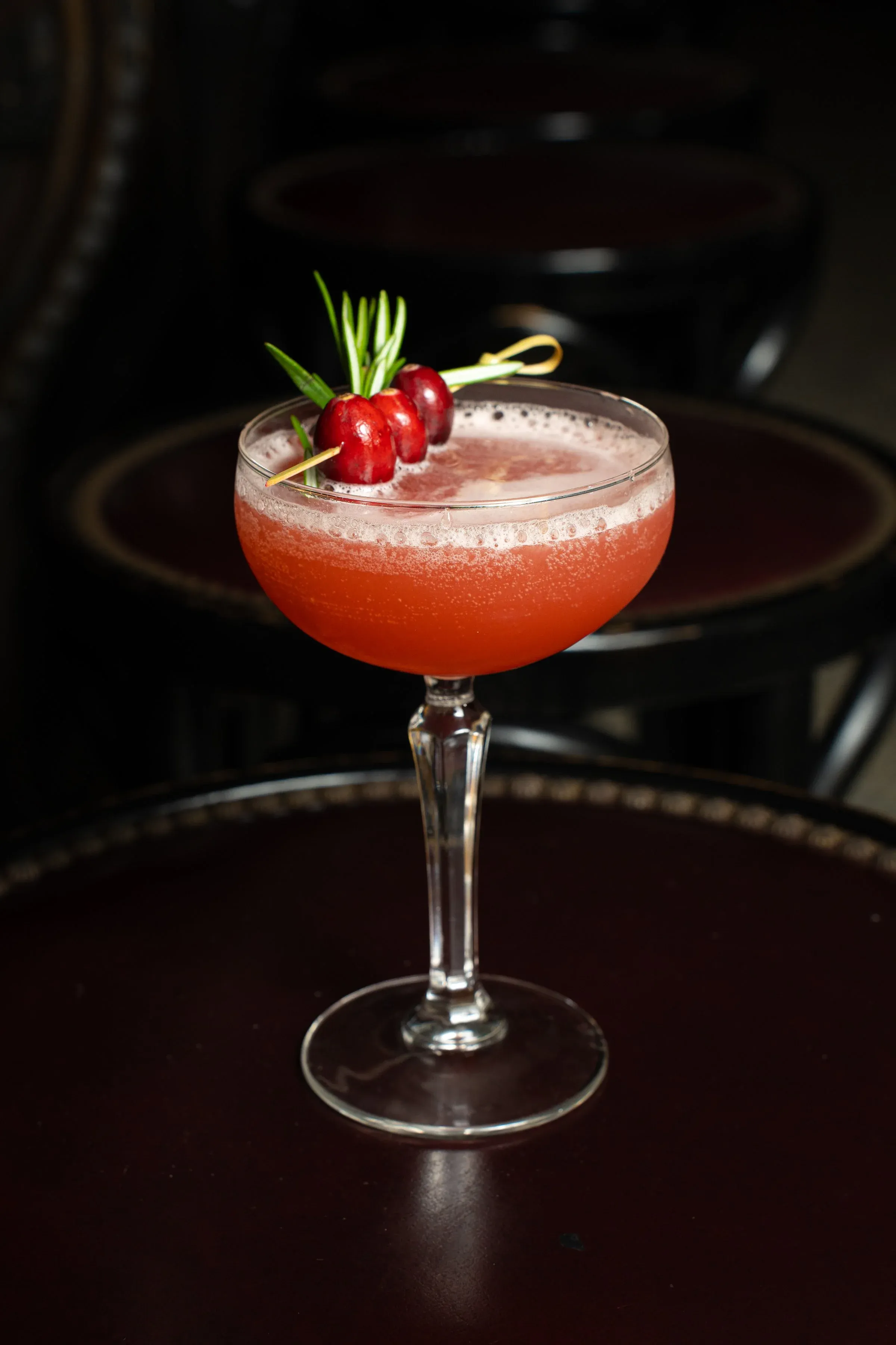 A red cocktail in a coupe glass garnished with cranberries and rosemary.