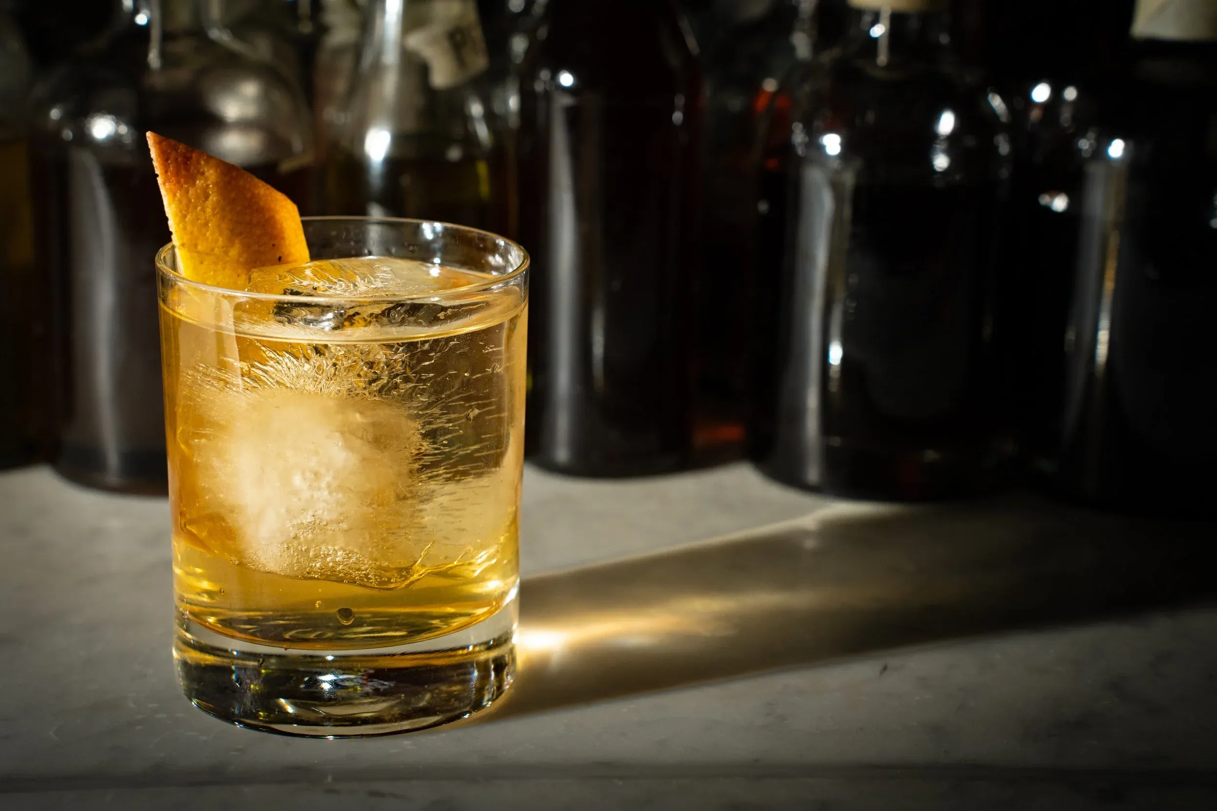 A golden cocktail with ice and an orange peel, surrounded by dark bottles.