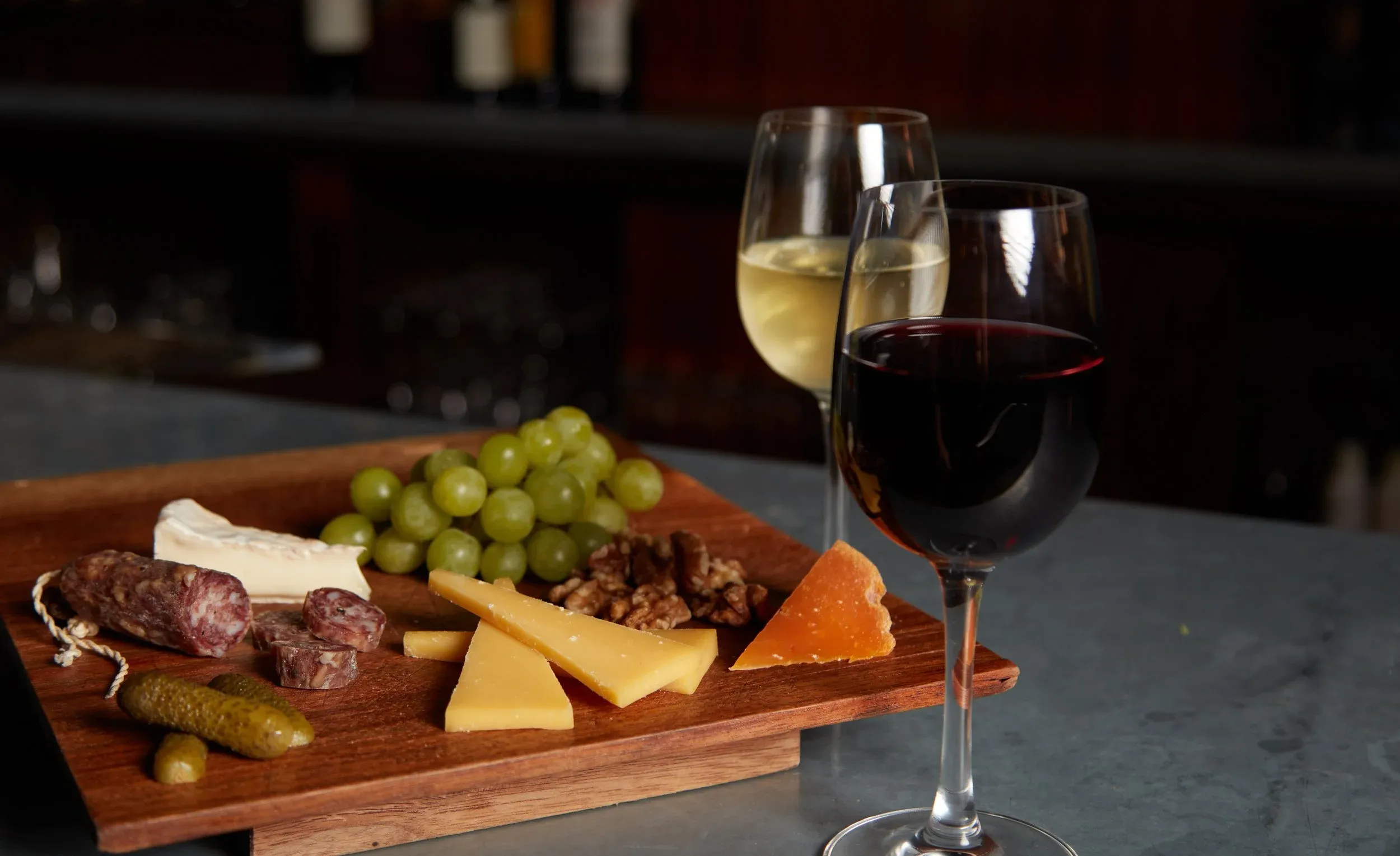 A wooden board with cheese, grapes, meats, and pickles next to two wine glasses.