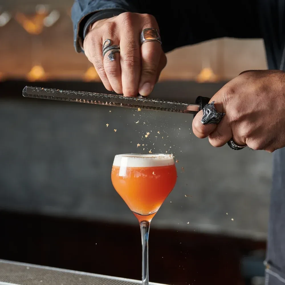 Hands grate garnish over a red cocktail in a coupe glass.