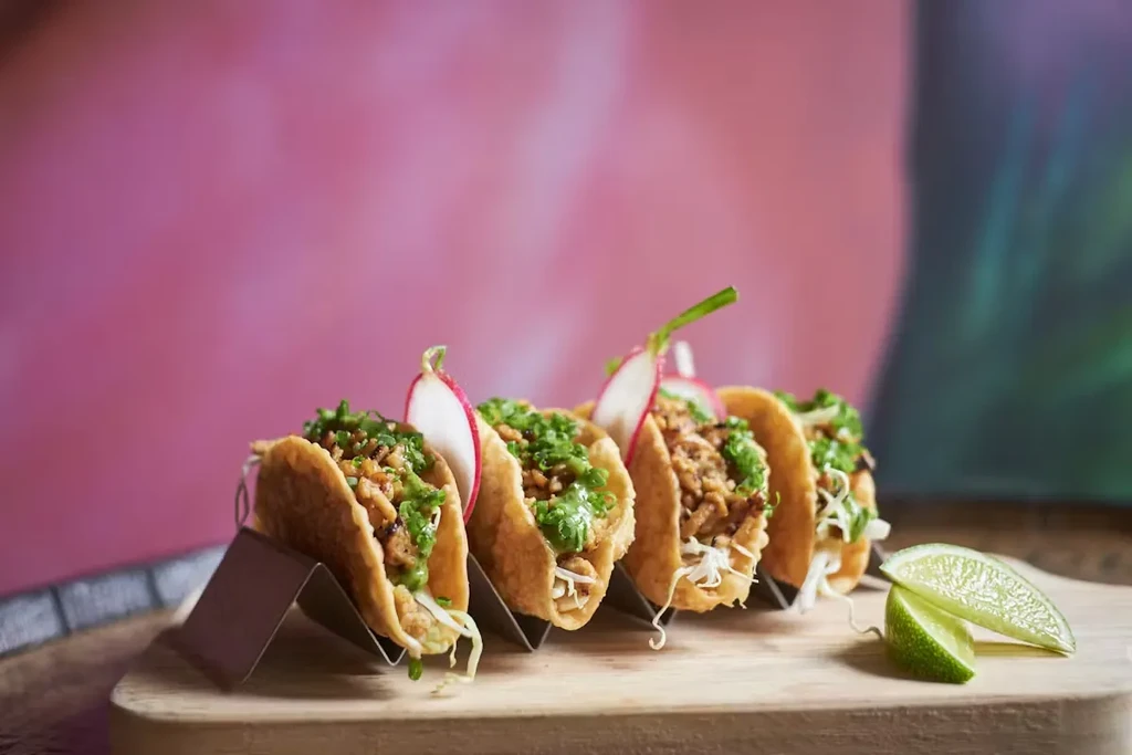 Five street tacos with radishes and lime wedges on a wooden board.