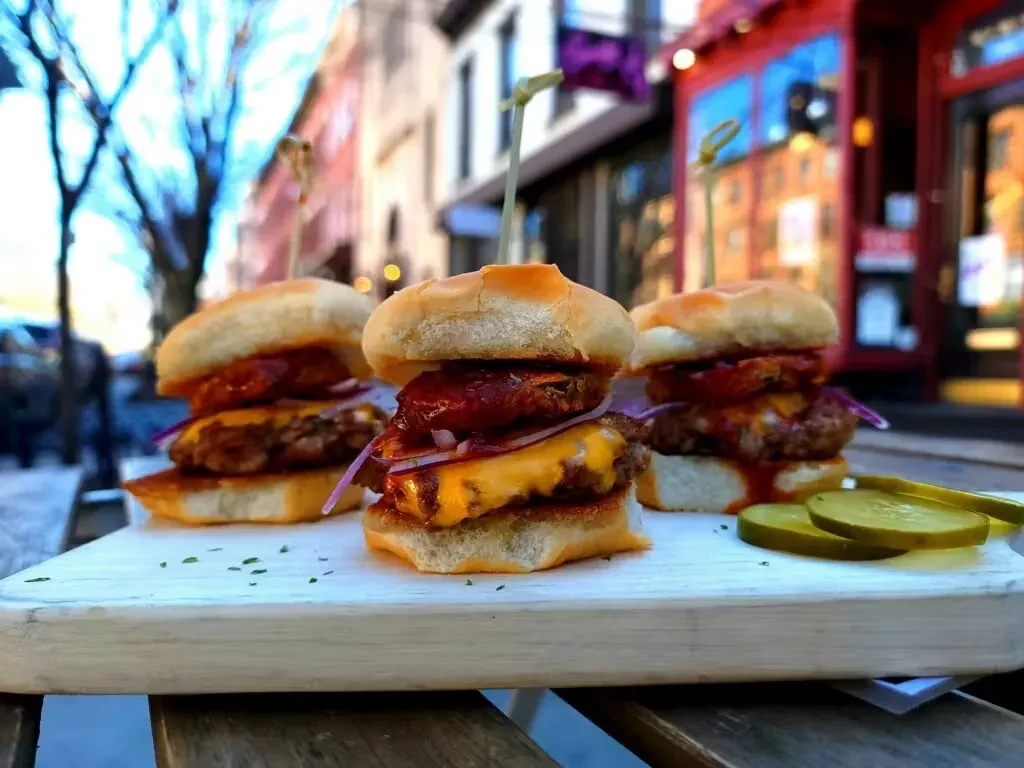 Three cheeseburger sliders with sauce, onions, and pickles on a board.
