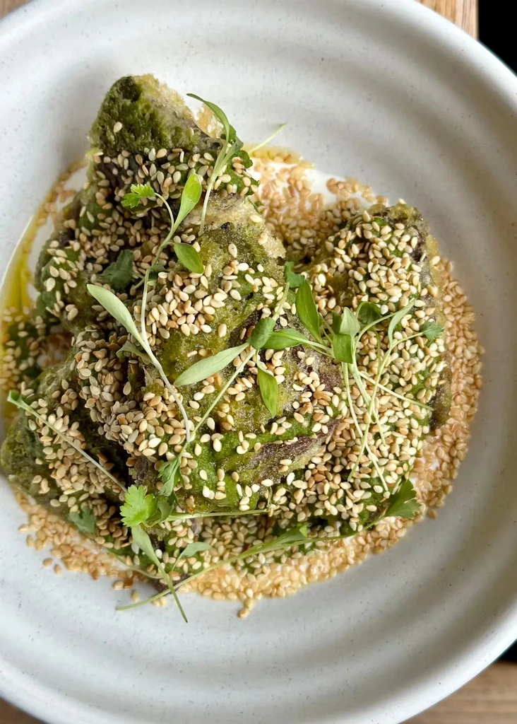 Green food, possibly tempura, covered in sesame seeds and fresh herbs.