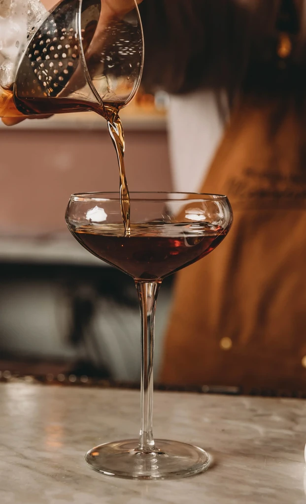 A bartender pours a dark cocktail into a coupe glass.