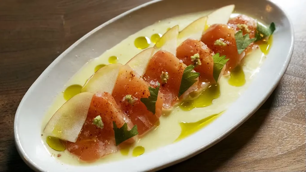 A white plate with sliced pears, salmon, herbs, and olive oil.