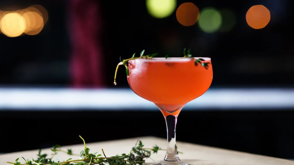 A coral-colored cocktail with a sprig of thyme and blurred lights in the background.