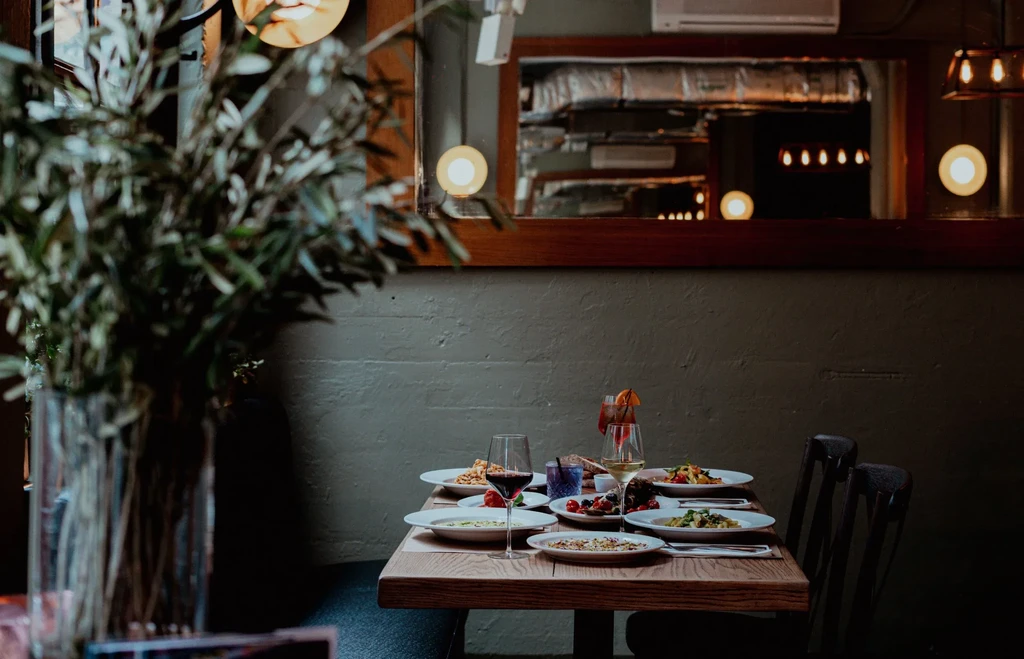 A candlelit restaurant table set with wine glasses, cocktails, and multiple dinner plates.