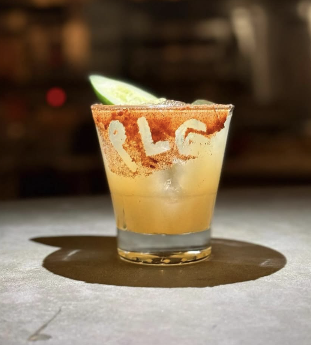A yellow cocktail with a spiced rim and cucumber slice on a table.