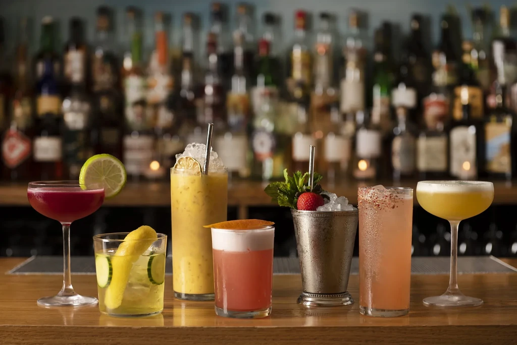 Seven colorful cocktails arranged on a bar counter, with liquor bottles blurred in the background.