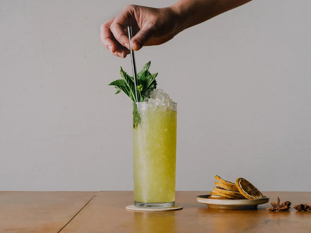 Hand inserting metal straw into tall green iced drink with mint.