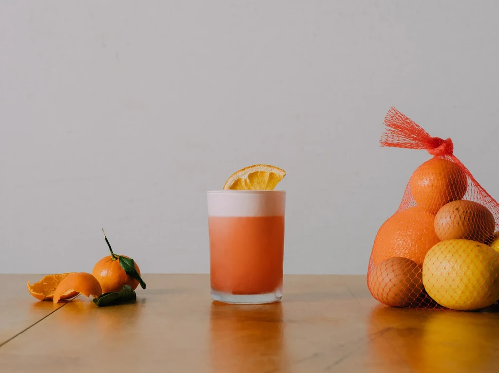 A cocktail and citrus fruits, some in a red mesh bag.