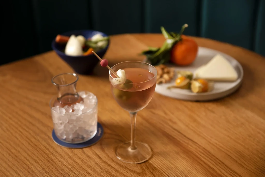 A pink cocktail with snacks, including cheese, fruits, and pickled vegetables.