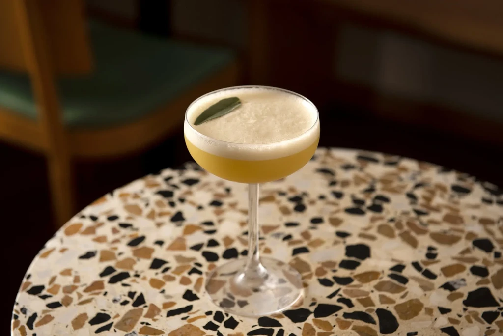A foamy yellow cocktail with a leaf garnish on a speckled table.