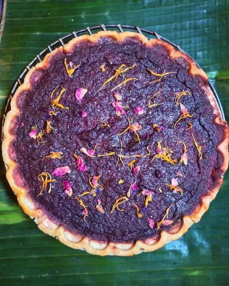 A baked purple pie with flower petals and zest on a banana leaf.