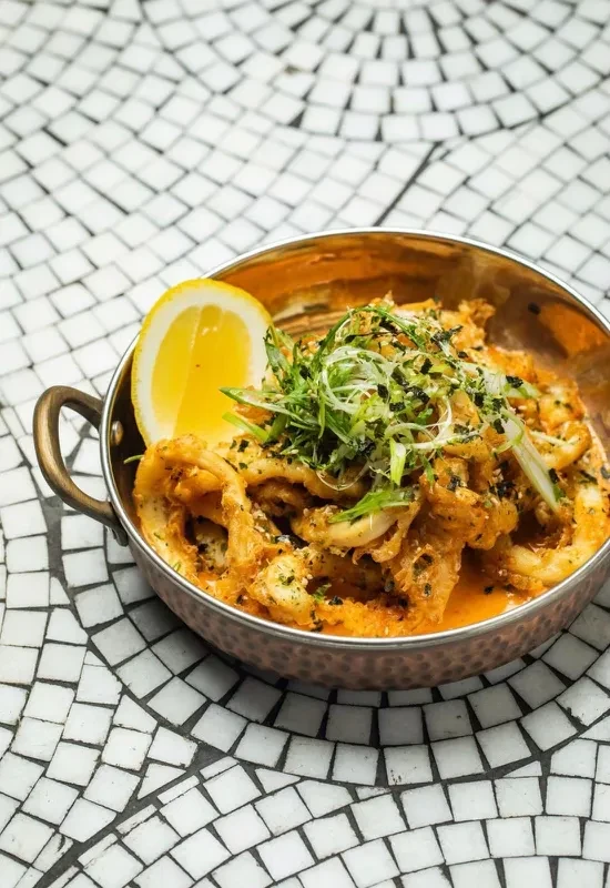A bowl of calamari with a lemon wedge and scallions on a mosaic table.