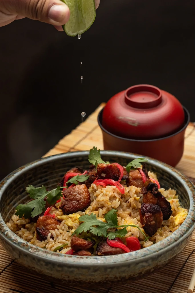 A hand squeezes a lime over fried rice with pork belly and cilantro.