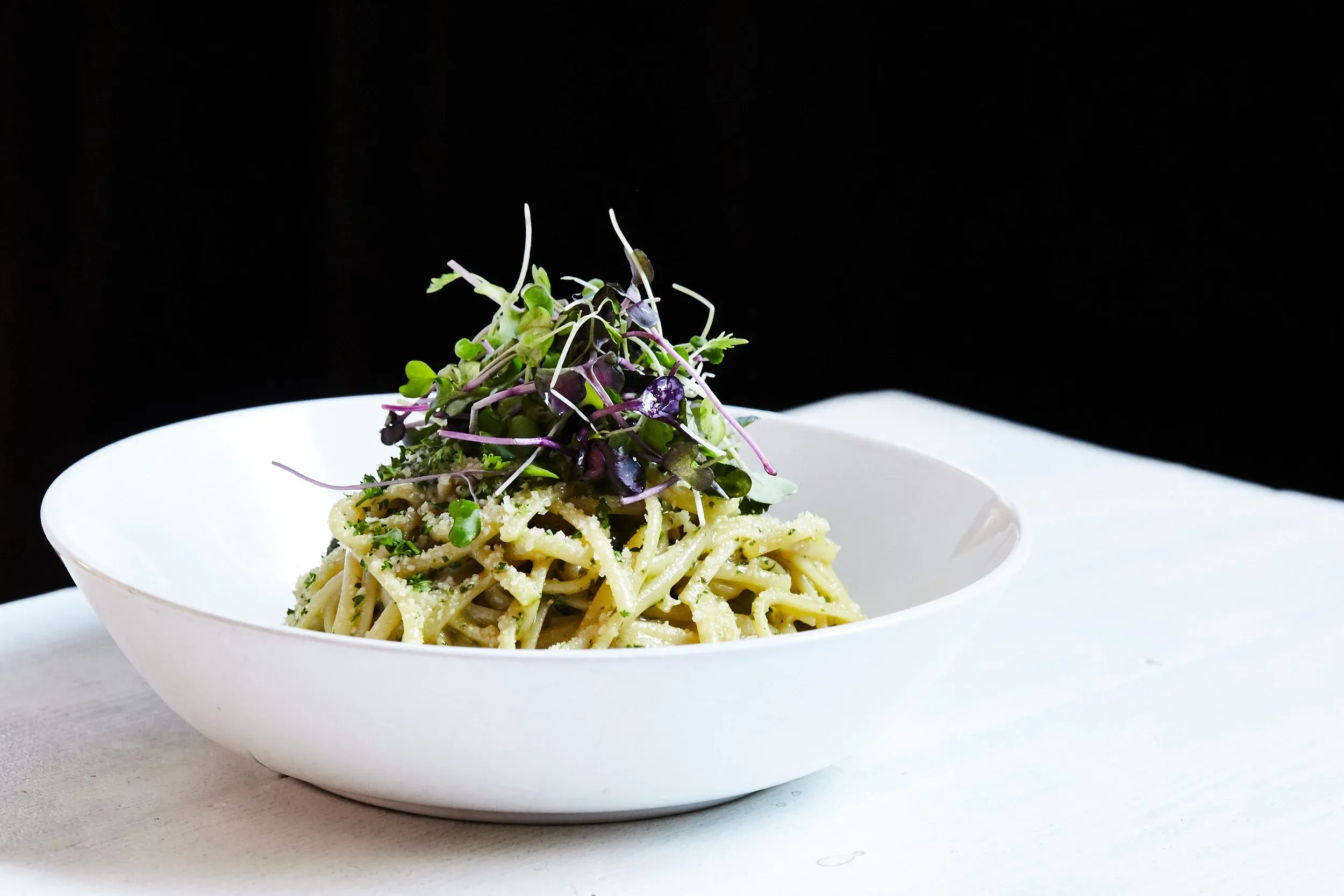 A white bowl filled with pasta, green sauce, cheese, and microgreens.