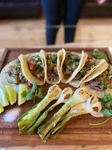 Five tacos, grilled onions, limes, and cilantro on a wooden board.