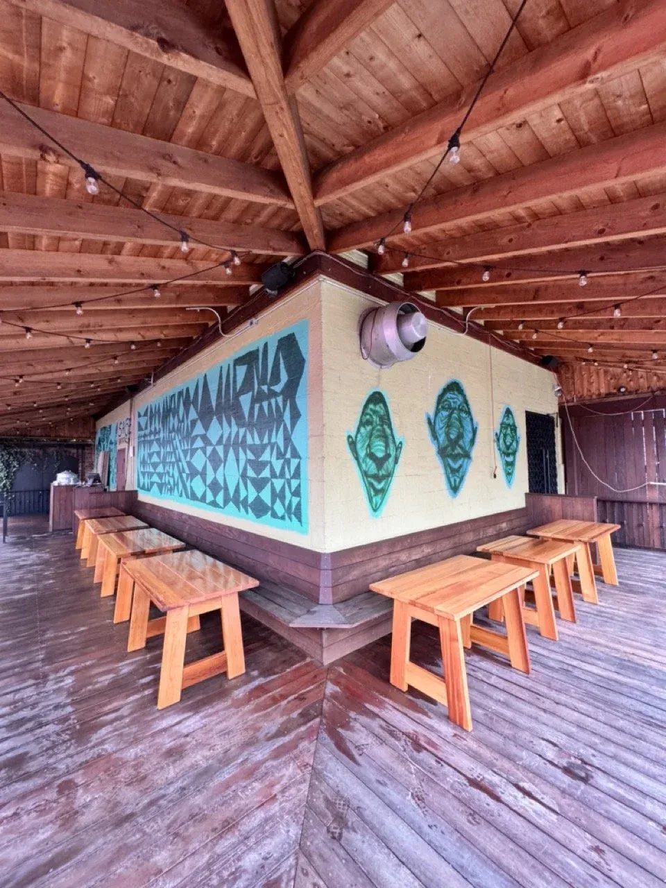 An outdoor patio with wood tables, a colorful mural, and string lights.