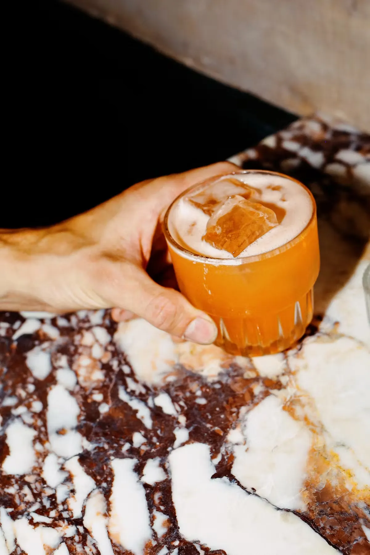 Hand holding an orange cocktail with ice on a marble table.