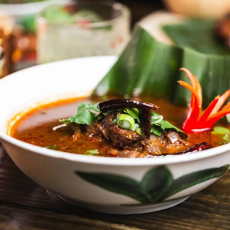 A spicy soup with meat, cilantro, and chili peppers in a white bowl.