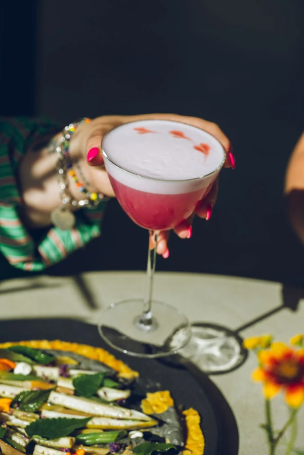 A person holding a pink cocktail with white foam and small hearts.