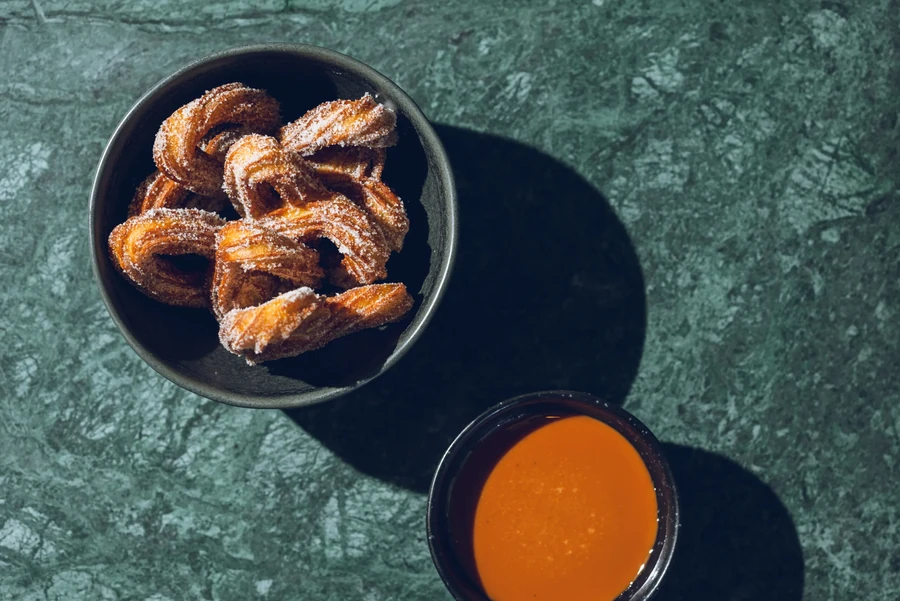 Sugar-dusted churros in a bowl, with a side of orange dipping sauce.