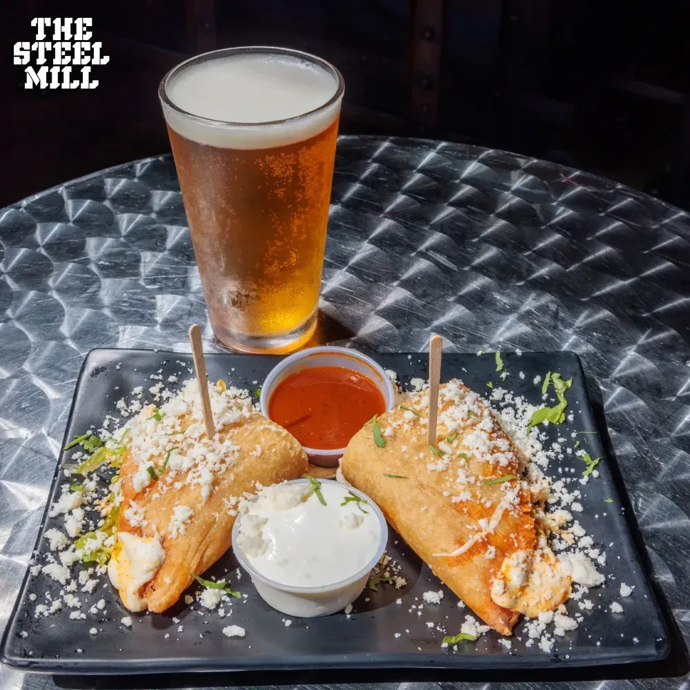 Two fried tacos and two dipping sauces with a beer on a metallic table.