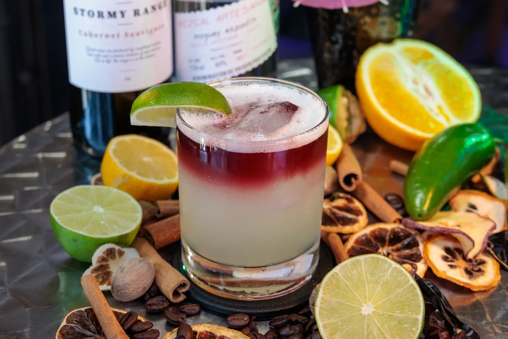 A layered red and white cocktail with a lime garnish, surrounded by fruit.