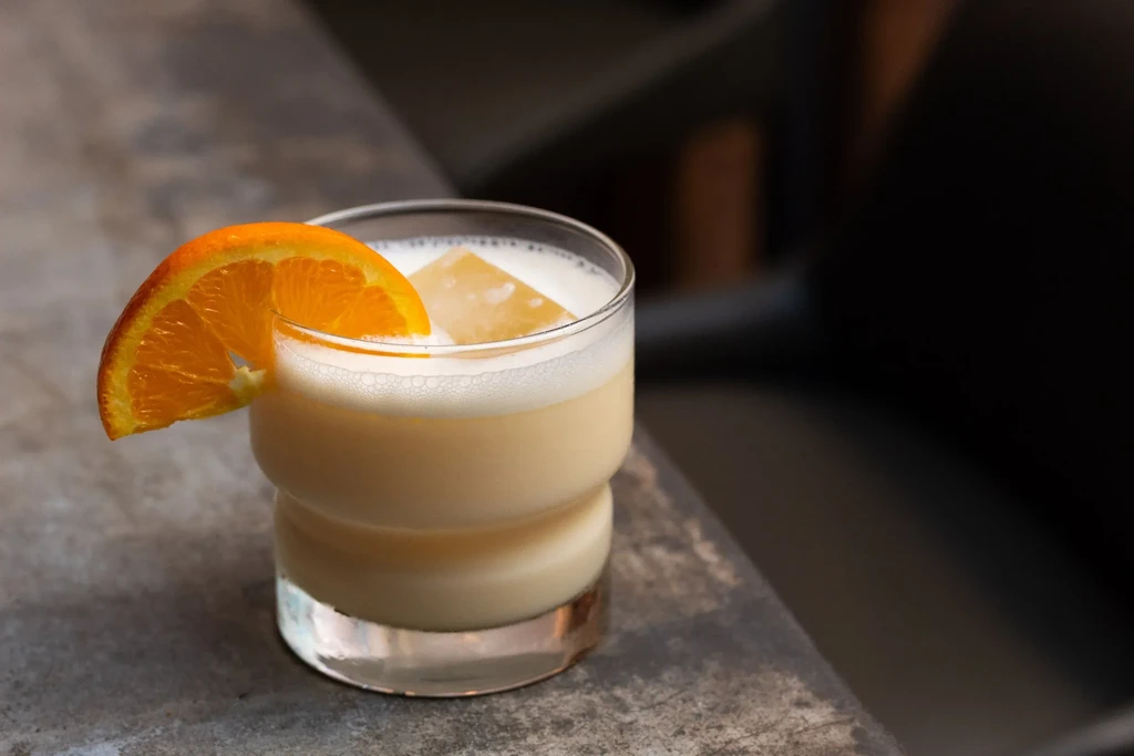 A creamy cocktail with an orange slice and ice cube on a gray surface.