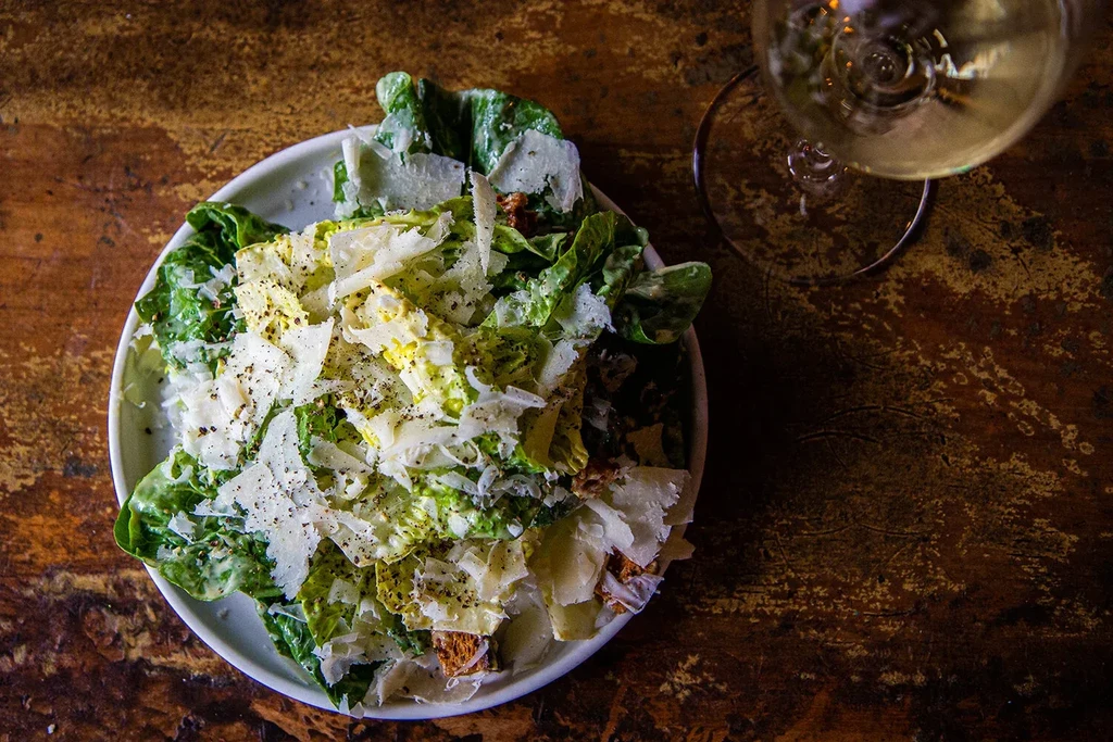 Caesar salad with shaved parmesan and wine on a dark wooden table.