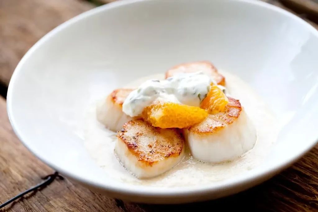 Seared scallops with orange segments and a creamy sauce in a white bowl.