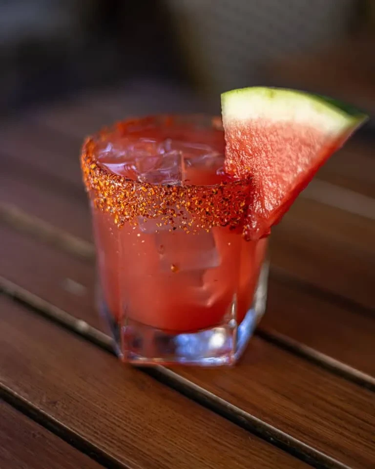 A red drink with ice, chili rim, and watermelon slice.