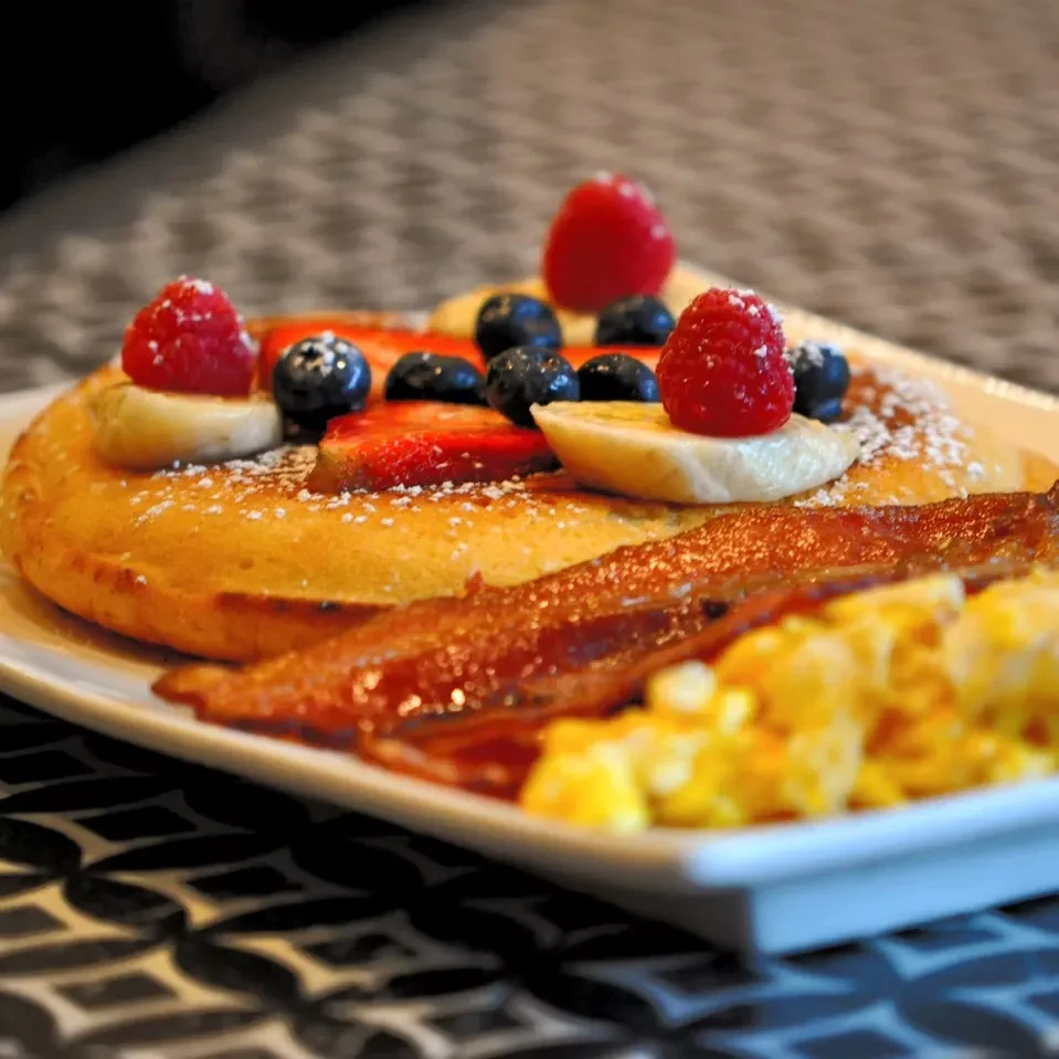 Pancakes with berries, bananas, bacon, and scrambled eggs on a white plate.
