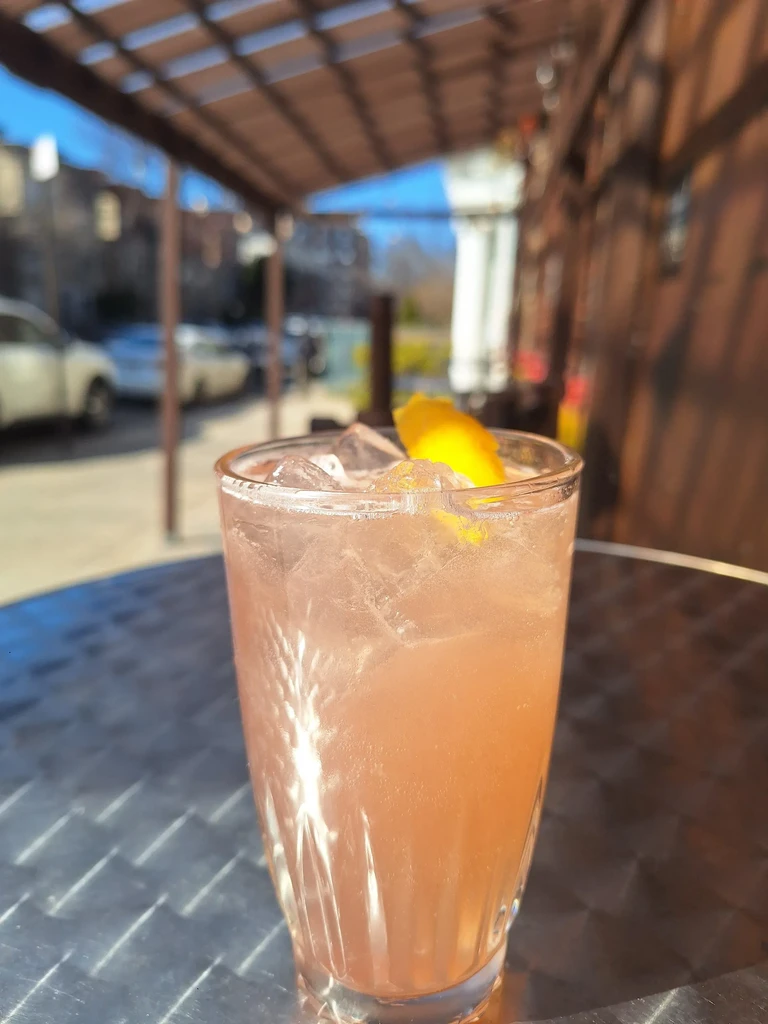 A pink cocktail with ice and a lemon twist on an outdoor table.