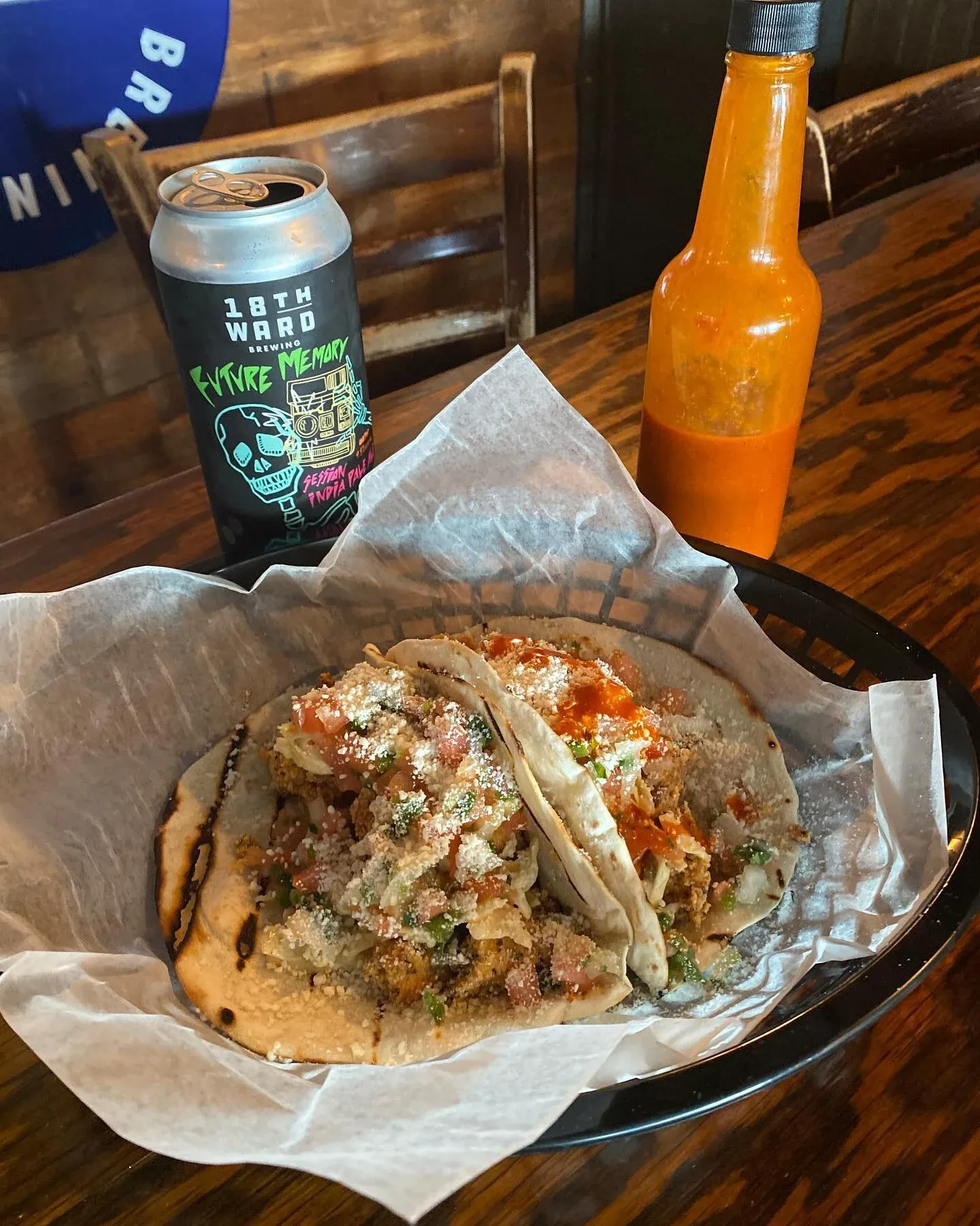 Two tacos, a beer can, and a hot sauce bottle on a wooden table.