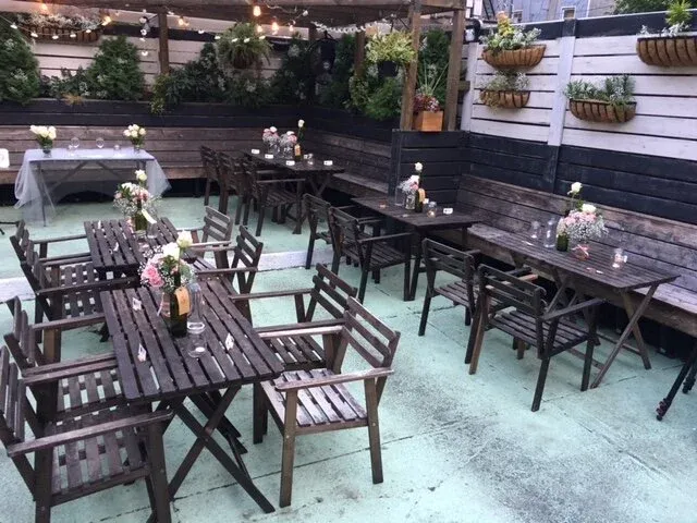 Outdoor patio with dark wooden tables, chairs, flowers, and string lights.