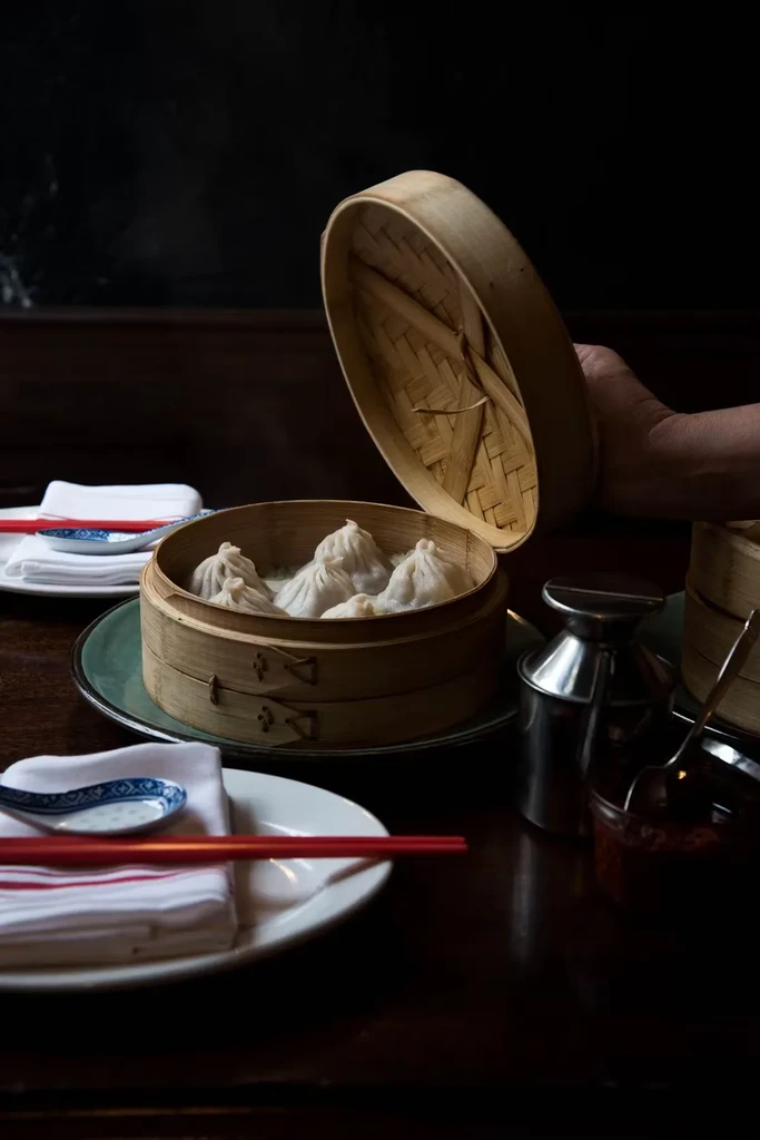 A hand lifts the lid off a bamboo steamer of dumplings.