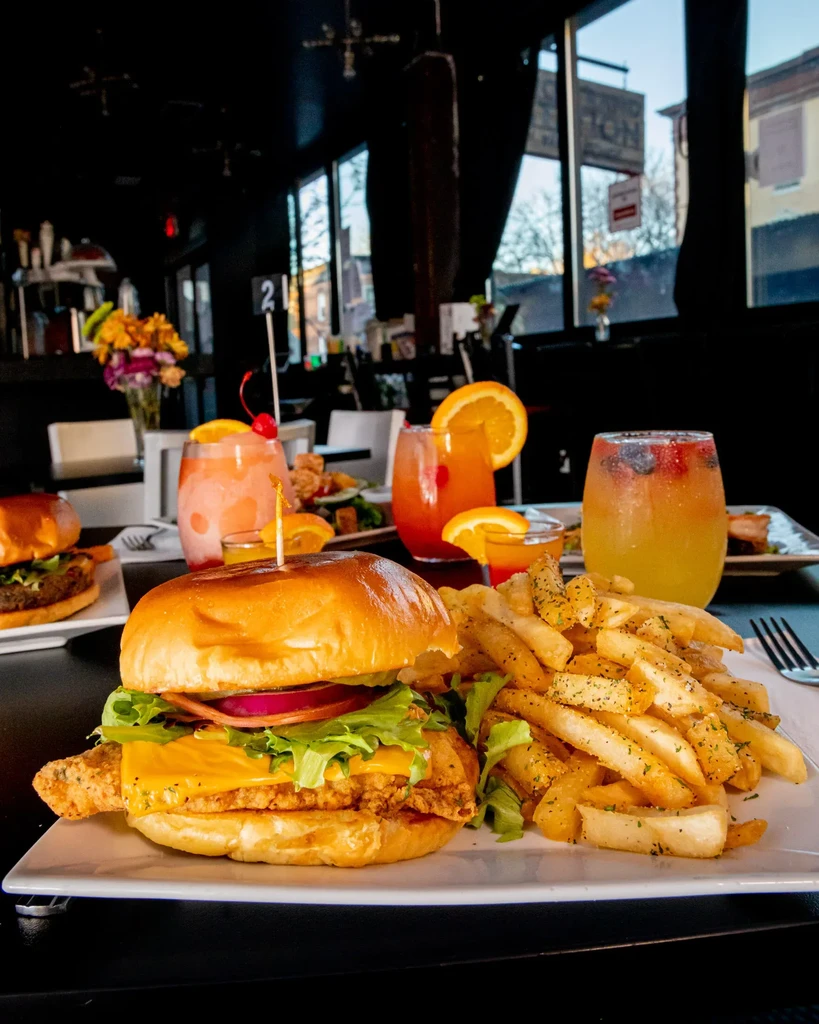 A fried fish sandwich with fries and three colorful cocktails on a table.
