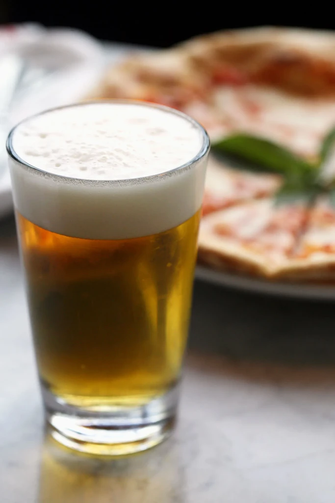 A glass of golden beer with foam next to a pizza.