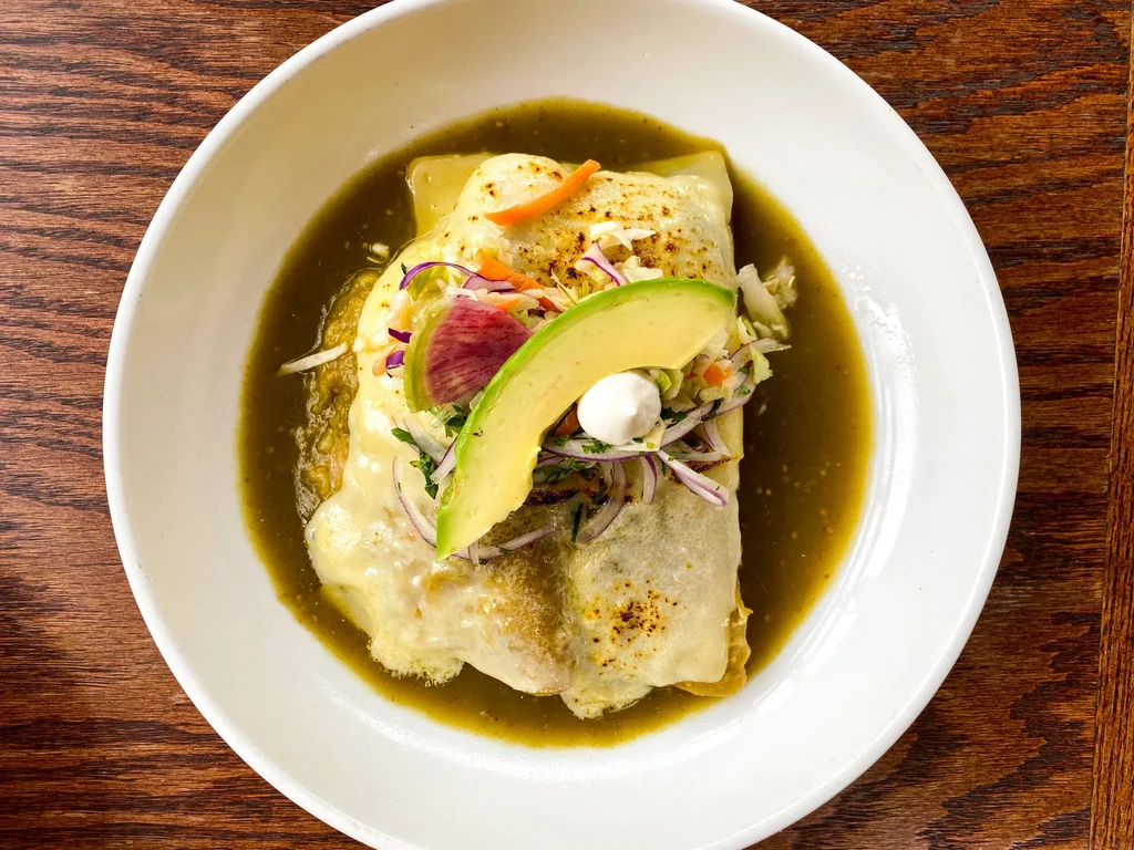 A large burrito in green sauce, topped with cheese, avocado, and vegetables.